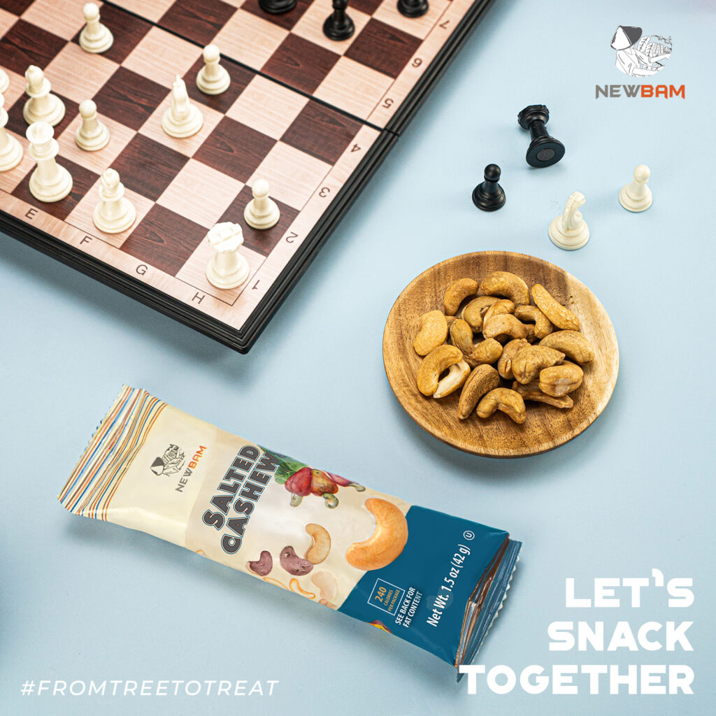NEWBAM Sea Salt cashew snack pack beside a wooden bowl of cashews and a chessboard.