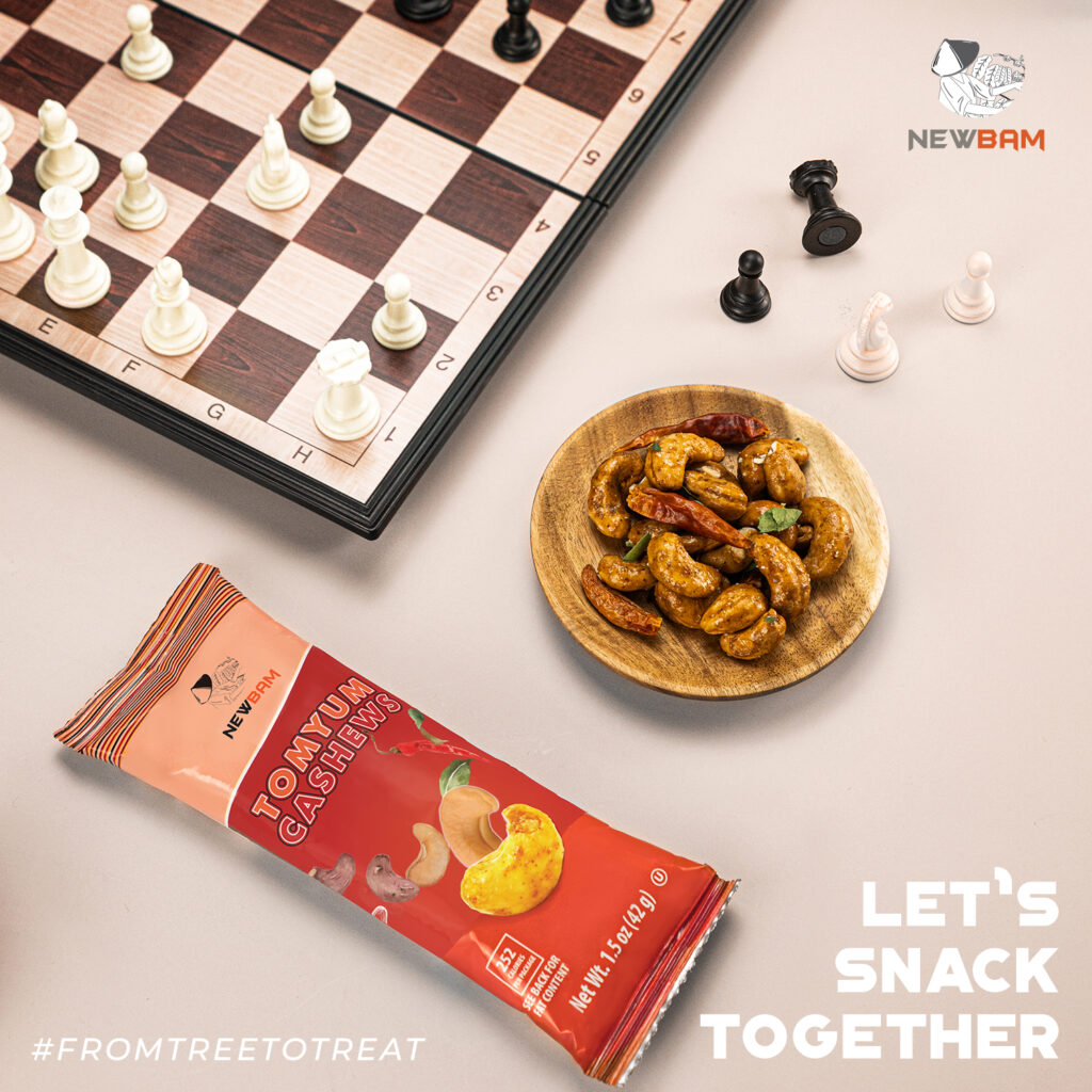 Tom Yum cashews in a wooden bowl beside a chessboard and a NEWBAM Tom Yum pouch.
