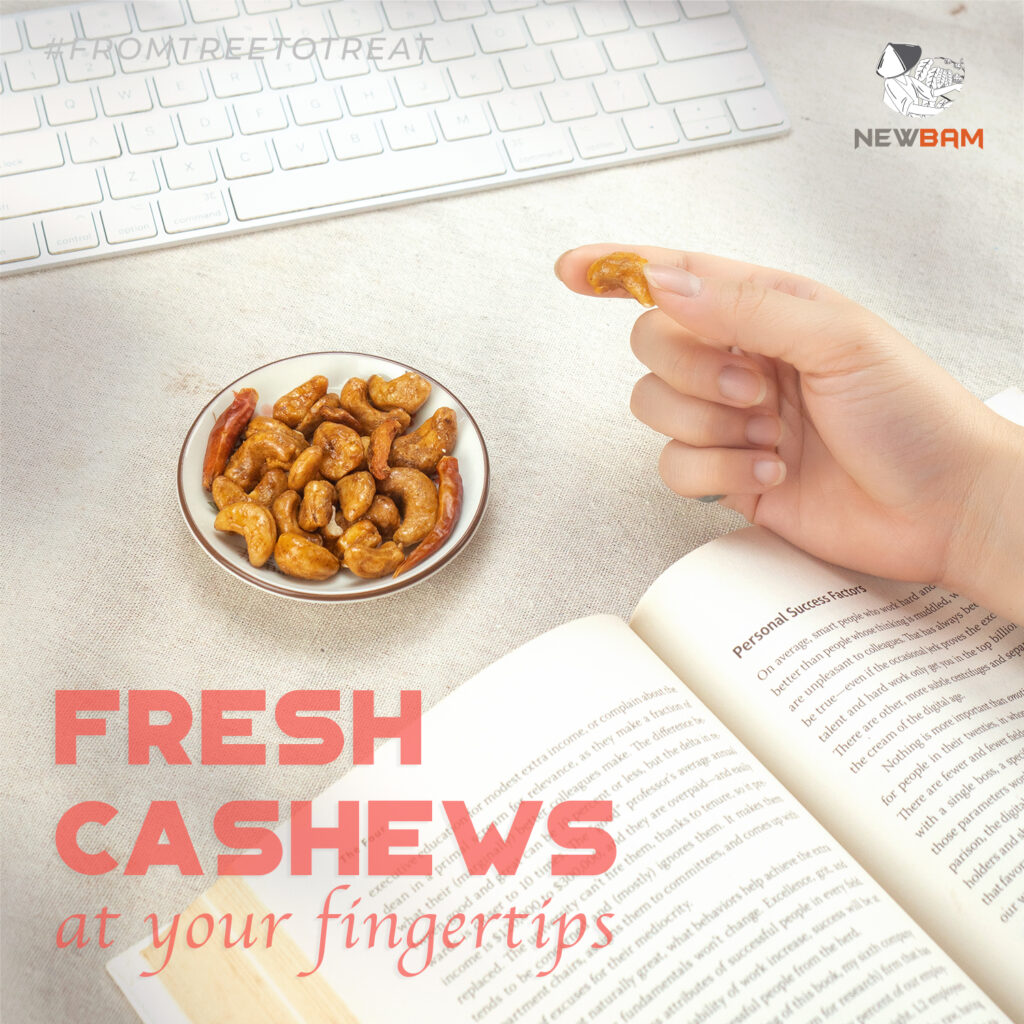 Tom Yum cashews in a bowl and a hand holding a cashew beside a keyboard and an open book.